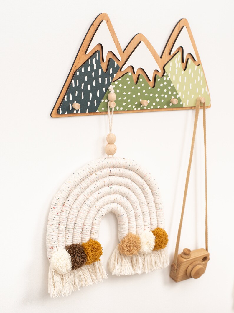 May include: A wooden mountain-shaped wall hook with three hooks and a white and green painted design. A white and brown macrame rainbow with pom-poms hangs from one of the hooks. A wooden camera hangs from another hook.