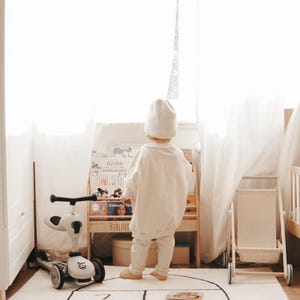 May include: A child wearing a white beanie, sweatshirt, and pants stands before a wooden bookshelf filled with books. To the left is a white three-wheeled scooter, and to the right is a small wooden cart. A hopscotch rug with numbers lies on the floor.