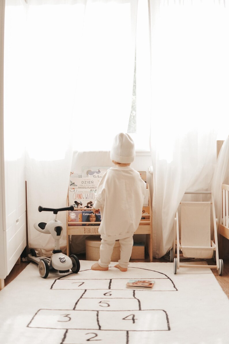 May include: A child wearing a white beanie, sweatshirt, and pants stands before a wooden bookshelf filled with books. To the left is a white three-wheeled scooter, and to the right is a small wooden cart. A hopscotch rug with numbers lies on the floor.