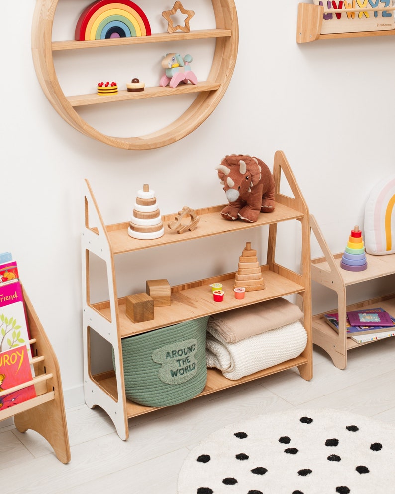 May include: A white and wood three-tiered shelf unit with a green storage basket labeled "Around the World". The shelf unit is filled with toys, books, and blankets. The shelf unit is in front of a white wall with a round wooden shelf on the wall above it.