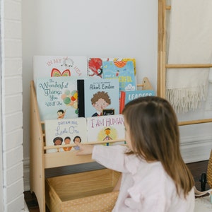 May include: A wooden bookshelf with a storage bin at the bottom. The bookshelf is filled with colorful children's books with titles like "The Wonderful Things You Will Be", "Dream Big, Little One", and "I Am Human". A young child is reaching for a book on the top shelf.