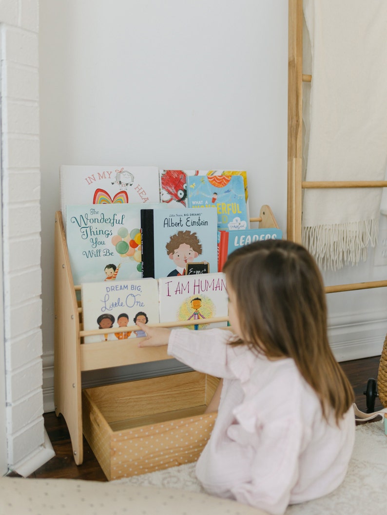 May include: A wooden bookshelf with a storage bin at the bottom. The bookshelf is filled with colorful children's books with titles like "The Wonderful Things You Will Be", "Dream Big, Little One", and "I Am Human". A young child is reaching for a book on the top shelf.