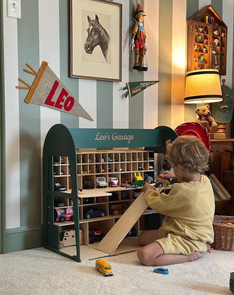 K&ouml;nnte beinhalten: Ein dunkelgr&uuml;nes Holzspielzeug-Garage mit mehreren F&auml;chern, die mit Spielzeugautos gef&uuml;llt sind. Die Garage hat eine Rampe und den Text "Leo's Garage" oben. Ein Kind spielt mit den Spielzeugautos. Der Raum hat gestreifte Tapeten und dekorative Wimpel.