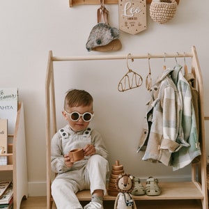 May include: A wooden clothing rack with a child sitting on the bottom shelf. The child is wearing a light brown shirt, overalls, white sneakers, and sunglasses. There are two shirts hanging on the rack, a plaid shirt and a light blue shirt. A wooden toy lion is sitting on the shelf next to the child. The rack has a shelf on top and a shelf on the bottom. There is a wooden name sign above the rack that says "Leo".