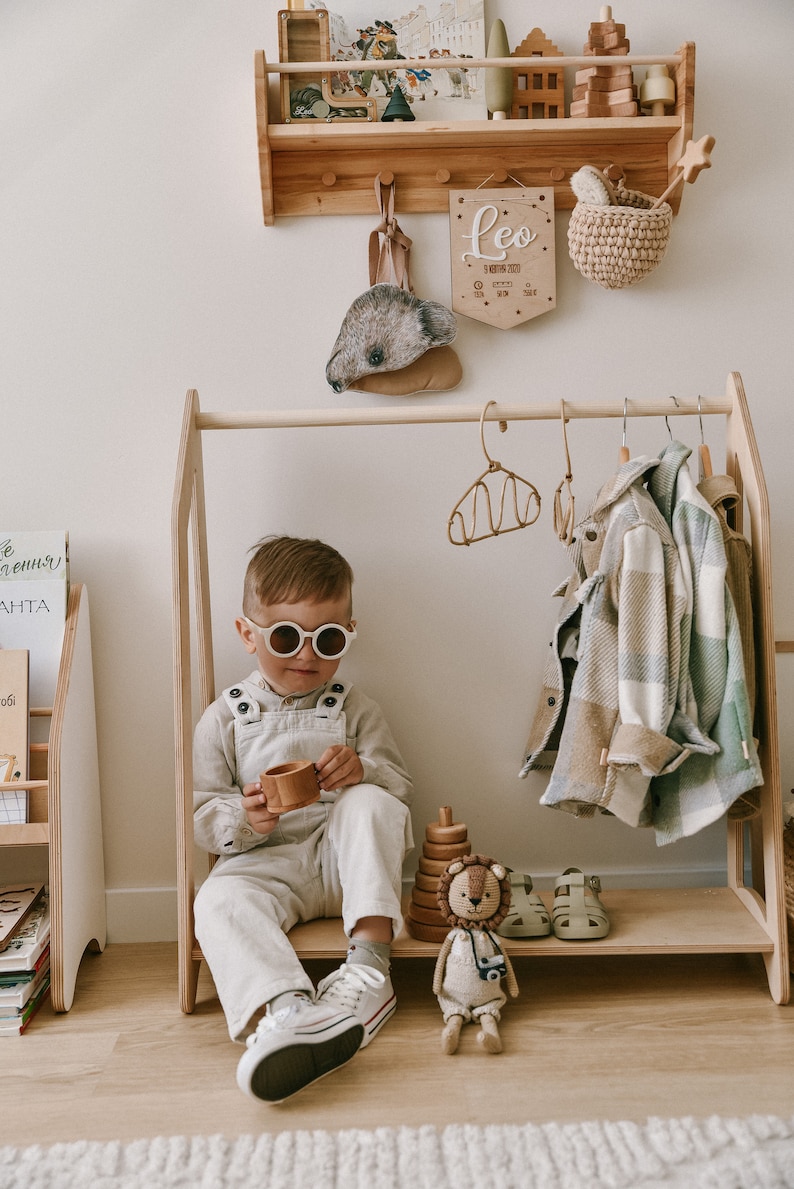 May include: A wooden clothing rack with a child sitting on the bottom shelf. The child is wearing a light brown shirt, overalls, white sneakers, and sunglasses. There are two shirts hanging on the rack, a plaid shirt and a light blue shirt. A wooden toy lion is sitting on the shelf next to the child. The rack has a shelf on top and a shelf on the bottom. There is a wooden name sign above the rack that says "Leo".