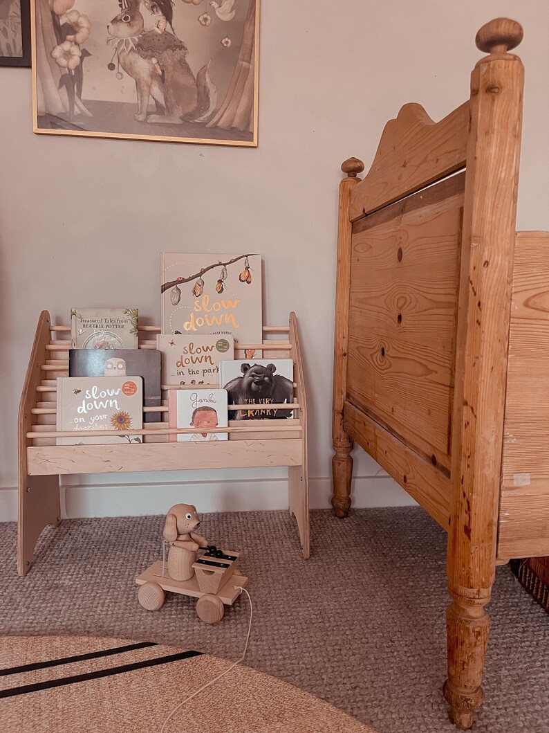 May include: A light-colored wooden bookshelf holding several children's books with titles including Slow Down, The Very Cranky Bear, and Treasured Tales from Beatrix Potter. A wooden pull-toy dog with a xylophone sits on the floor in front of the shelf.