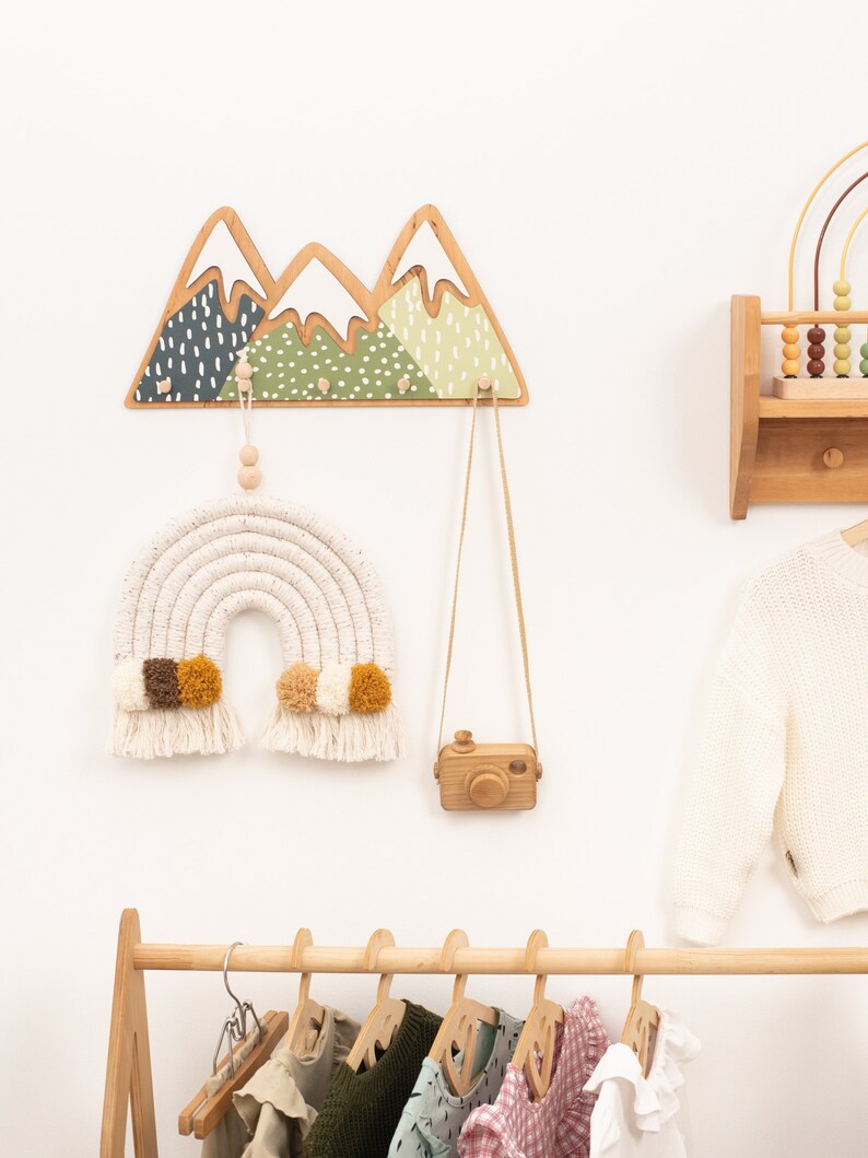 May include: Wooden mountain-shaped wall hooks with green and white paint, a rainbow macrame wall hanging with brown and yellow pom-poms, a wooden camera, and a wooden clothes rack with hangers and children's clothing.