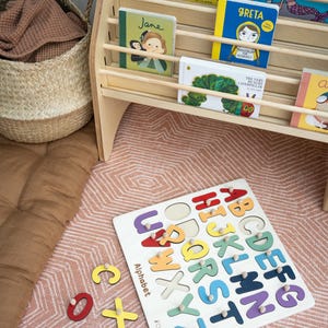 May include: A wooden alphabet puzzle board with colorful letters, featuring the text 'Alphabet' and 'Childuniverse'. Beside it, a wooden bookshelf holds children's books including 'Artists', 'That's not my mermaid', 'Jane', 'Greta', and 'The Very Hungry Caterpillar'.