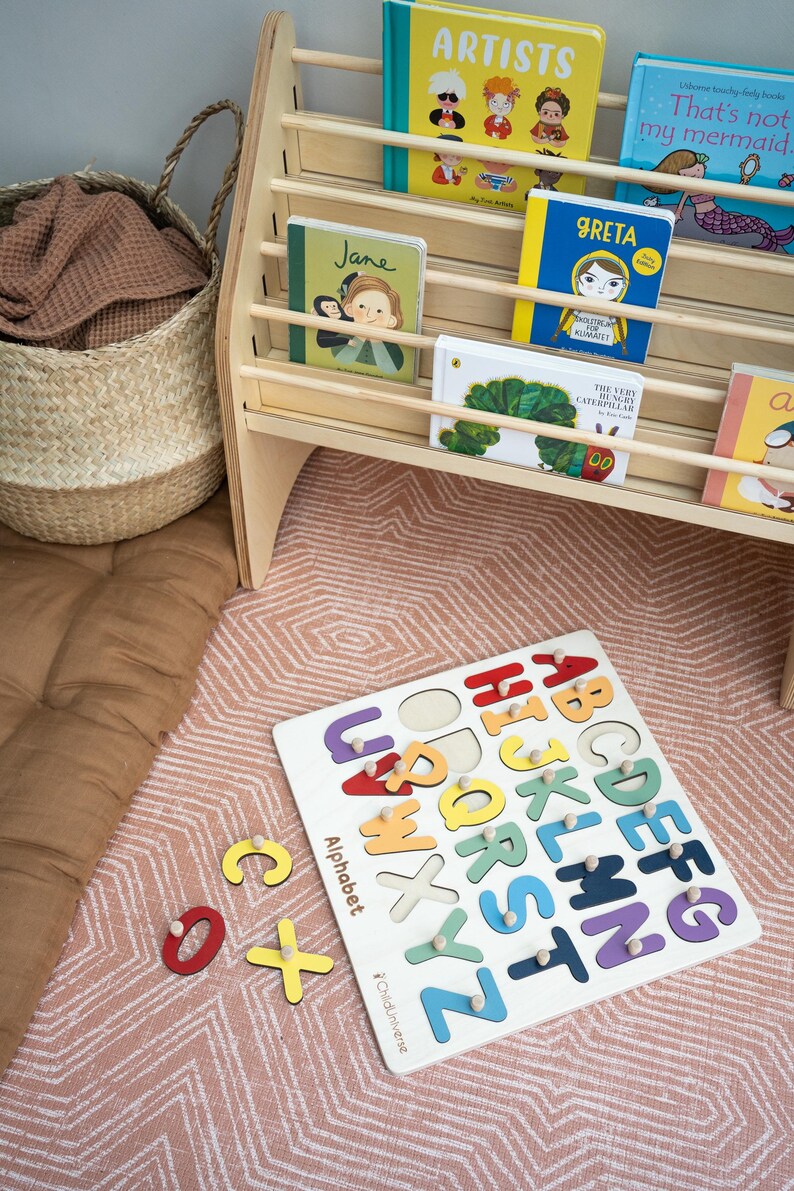 May include: A wooden alphabet puzzle board with colorful letters, featuring the text 'Alphabet' and 'Childuniverse'. Beside it, a wooden bookshelf holds children's books including 'Artists', 'That's not my mermaid', 'Jane', 'Greta', and 'The Very Hungry Caterpillar'.