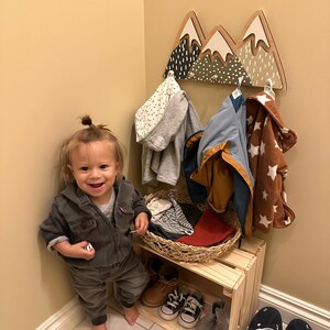 May include: A toddler stands in front of a wooden mountain-shaped coat rack with a basket of clothes underneath. The rack has three peaks painted in green, white, and black with white polka dots. The toddler is wearing a dark denim jumpsuit and has a small bun in their hair.