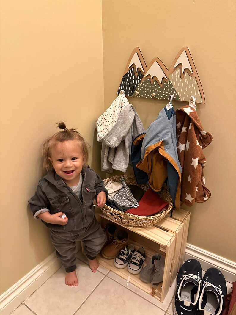 May include: A toddler stands in front of a wooden mountain-shaped coat rack with a basket of clothes underneath. The rack has three peaks painted in green, white, and black with white polka dots. The toddler is wearing a dark denim jumpsuit and has a small bun in their hair.