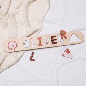 May include: Wooden name puzzle spelling "OLIVER" with various colors and patterns. The puzzle includes a compass design and a mountain-shaped piece. The letters are in different shades of brown and white. The puzzle is on a white surface.