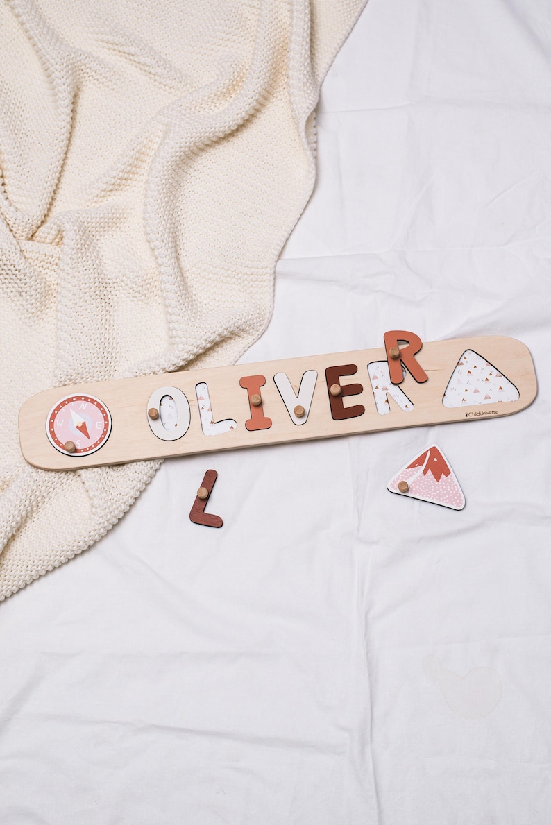 May include: Wooden name puzzle spelling "OLIVER" with various colors and patterns. The puzzle includes a compass design and a mountain-shaped piece. The letters are in different shades of brown and white. The puzzle is on a white surface.
