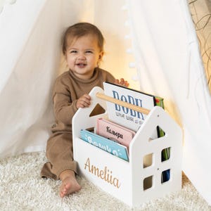 May include: A white wooden house-shaped bookshelf with a wooden handle, holding several colorful books. The name "Ameliia" is written in gold script on the front. The bookshelf is on a white rug.