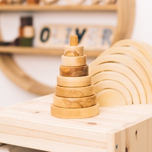 Könnte beinhalten: Ein Holz-Stapelspielzeug mit sieben Ringen in verschiedenen Größen, die in Pyramidenform angeordnet sind. Das Spielzeug ist aus Naturholz gefertigt und hat eine hellbraune Farbe. Das Spielzeug steht auf einem Holzregal mit dem Namen "OLIVER", der in Holzbuchstaben geschrieben ist.