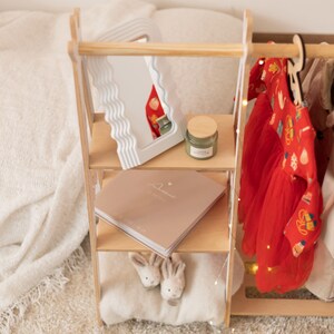 May include: A wooden doll clothes rack with three shelves. The top shelf has a white wavy mirror, the middle shelf has a book with the word "Dearest" on the cover, and the bottom shelf has a white pillow with two bunny-shaped stuffed animals. A red dress with a floral pattern hangs on a hanger on the right side of the rack.