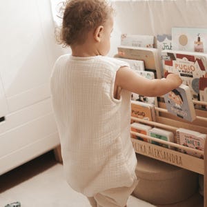 May include: A sleeveless, cream-colored muslin sleeping bag worn over light-colored leggings. The garment features a textured, crinkled fabric finish and a simple, loose-fitting design that allows for movement while standing near a wooden bookshelf.