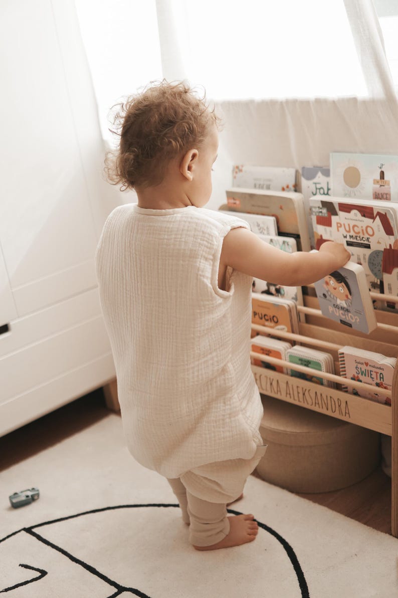 May include: A sleeveless, cream-colored muslin sleeping bag worn over light-colored leggings. The garment features a textured, crinkled fabric finish and a simple, loose-fitting design that allows for movement while standing near a wooden bookshelf.