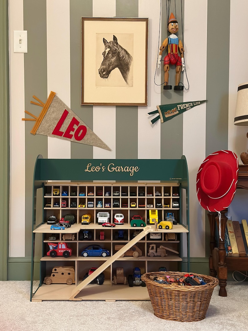K&ouml;nnte beinhalten: Ein Holzregal f&uuml;r Spielzeugautos mit der Aufschrift "Leo's Garage", gef&uuml;llt mit Miniatur-Spielzeugautos. Ein roter Cowboyhut h&auml;ngt in der N&auml;he. Ein gerahmtes Pferdeportr&auml;t und eine Pinocchio-Puppe sind ebenfalls zu sehen.