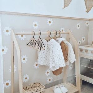 May include: Wooden children's clothing rack with a shelf, featuring a white and brown daisy pattern on the wall behind it. The rack has a wooden bar for hanging clothes and a shelf below for storage. There are two pairs of white shoes on the shelf and a woven basket.