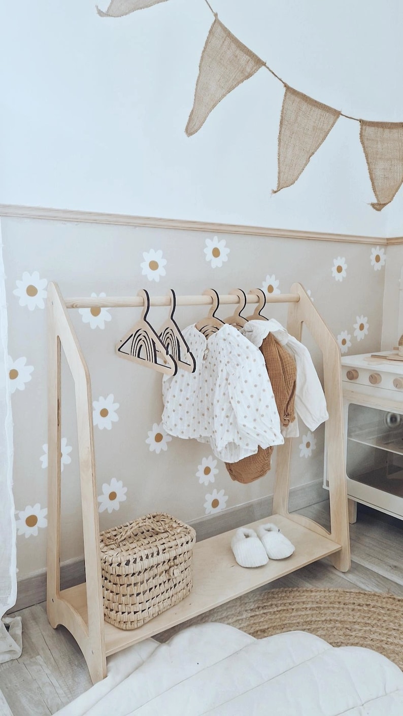 May include: Wooden children's clothing rack with a shelf, featuring a white and brown daisy pattern on the wall behind it. The rack has a wooden bar for hanging clothes and a shelf below for storage. There are two pairs of white shoes on the shelf and a woven basket.