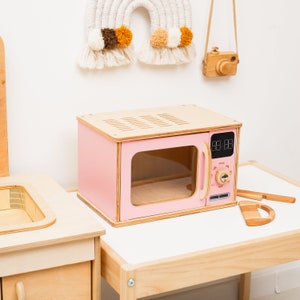 Può includere: Un forno a microonde giocattolo in legno rosa con una porta trasparente e un display digitale che mostra "00:00". Il forno a microonde è su un tavolo di legno.