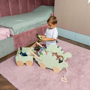 May include: A light green wooden pull-along toy wagon shaped like a dinosaur with a matching rectangular trailer. The dinosaur wagon features a long white pull cord with a handle, light wood wheels, and contains various small toy figures and books.