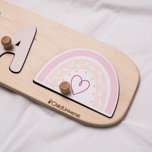 May include: Wooden puzzle with a light wood finish, featuring a pink and white rainbow design with a heart. The puzzle includes the letter "A" and two wooden knobs for grasping. The text "ChildUniverse" is printed on the puzzle.