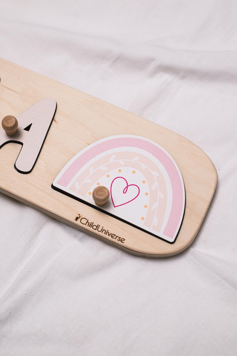 May include: Wooden puzzle with a light wood finish, featuring a pink and white rainbow design with a heart. The puzzle includes the letter "A" and two wooden knobs for grasping. The text "ChildUniverse" is printed on the puzzle.
