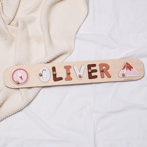 May include: Wooden name puzzle spelling "OLIVER" in various colors, with decorative images of a compass, a penguin, and a mountain. The puzzle is on a light-colored wooden board.