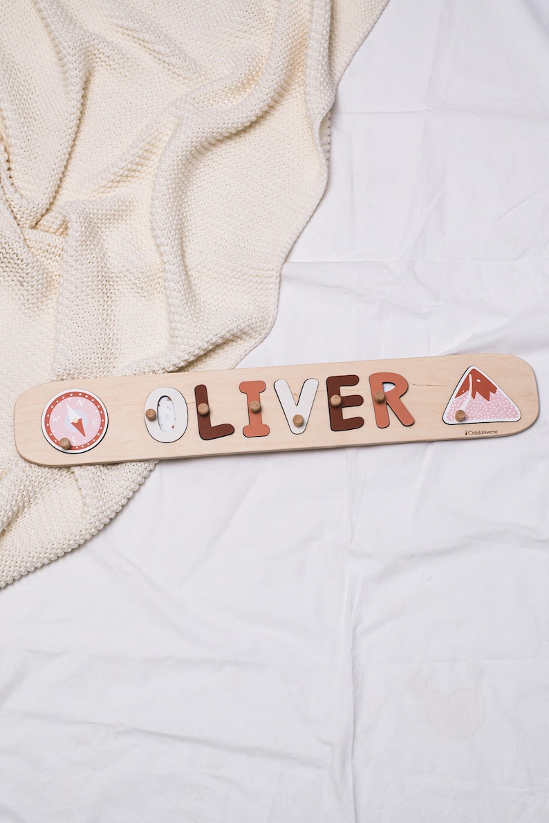 May include: Wooden name puzzle spelling "OLIVER" in various colors, with decorative images of a compass, a penguin, and a mountain. The puzzle is on a light-colored wooden board.