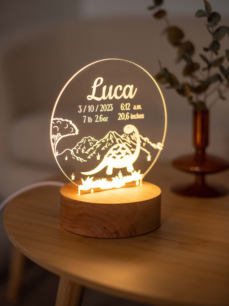 May include: A personalised night light with a dinosaur design. The clear acrylic light features the name "Luca", date, time, weight (3.2 kg), and height (52.3 cm). The light sits on a round wooden base and is illuminated.