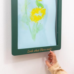 May include: A framed child's artwork showing a yellow flower and green leaves on a light blue background. The dark green frame has the text "Look what Oliver made!" in gold lettering.