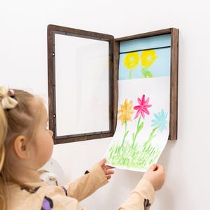 Op de afbeelding: Een donkerbruine houten displaylijst voor aan de muur, met een transparant voorpaneel dat openklapt om een opbergvak te onthullen. Binnenin wordt een wit vel papier met een handgeschilderde bloemenillustratie geplaatst of verwijderd.