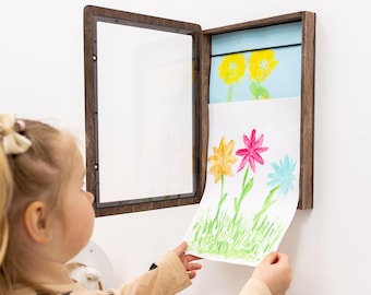 May include: A dark brown wooden hinged display frame mounted on a wall, featuring a clear front panel that opens to reveal a storage compartment. Inside, a white paper with a colorful hand-painted flower illustration is being placed or removed.