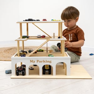 K&ouml;nnte beinhalten: Ein mehrst&ouml;ckiges Parkhaus aus Holz mit wei&szlig;em &Auml;u&szlig;eren und Akzenten aus Naturholz. Das Parkhaus hat mehrere Ebenen mit Rampen und Parkpl&auml;tzen, die mit Spielzeugautos gef&uuml;llt sind. Der Text "My Parking" ist auf der Vorderseite aufgedruckt.