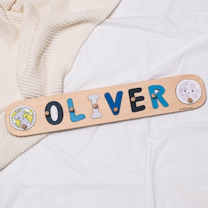 May include: Wooden name puzzle spelling "OLIVER" in blue, black, and gray letters. The puzzle has a light wood finish with a smiling moon and earth graphic on each end. A fun educational toy.