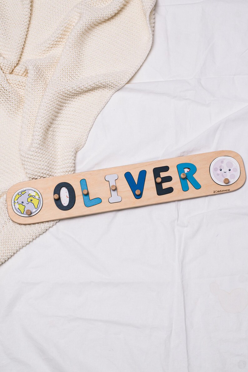 May include: Wooden name puzzle spelling "OLIVER" in blue, black, and gray letters. The puzzle has a light wood finish with a smiling moon and earth graphic on each end. A fun educational toy.