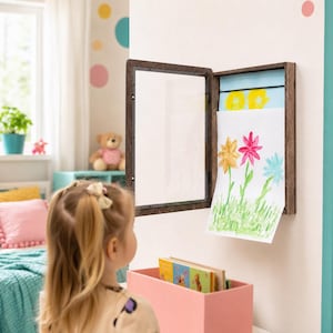 Może przedstawiać: Ciemnobrązowa drewniana ramka ścienna z otwieranymi szklanymi drzwiczkami, ukazująca białą kartkę papieru z ręcznie malowaną ilustracją kwiatów w kolorach żółtym, różowym i niebieskim, zamontowana na jasnej powierzchni ściany.