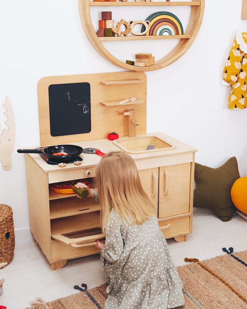 May include: A child plays with a wooden toy kitchen set. The kitchen features a stove with an oven, a sink, and a chalkboard. The child is wearing a green and white polka dot dress and is opening the oven door.