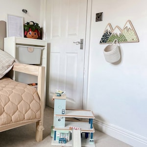 May include: A wooden toy car garage with a ramp and a helicopter on top. The garage is painted in shades of blue and green. The garage is sitting on a light brown carpet in a room with white walls.