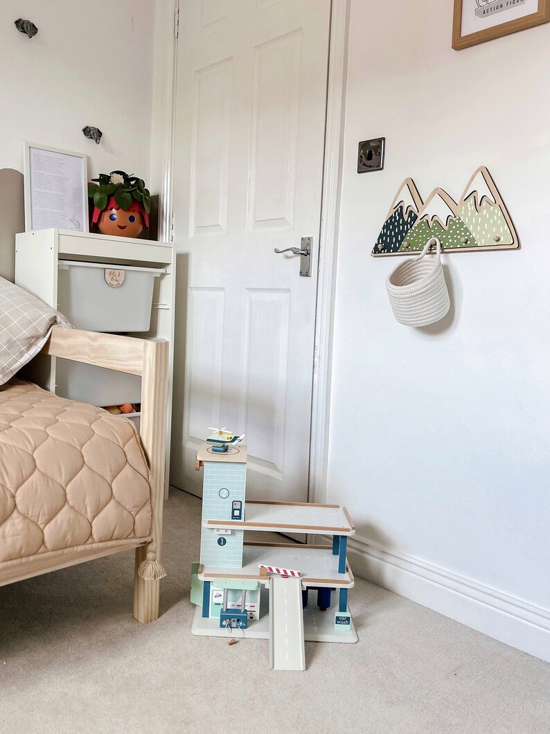 May include: A wooden toy car garage with a ramp and a helicopter on top. The garage is painted in shades of blue and green. The garage is sitting on a light brown carpet in a room with white walls.