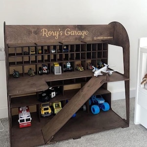 K&ouml;nnte beinhalten: Ein dunkelbraunes Spielzeug-Garage aus Holz mit mehreren F&auml;chern und einer Rampe. Es ist mit Spielzeugautos und -lastwagen gef&uuml;llt. Der Name "Rory's Garage" ist oben eingraviert. Ideal zur Aufbewahrung von Spielzeug.