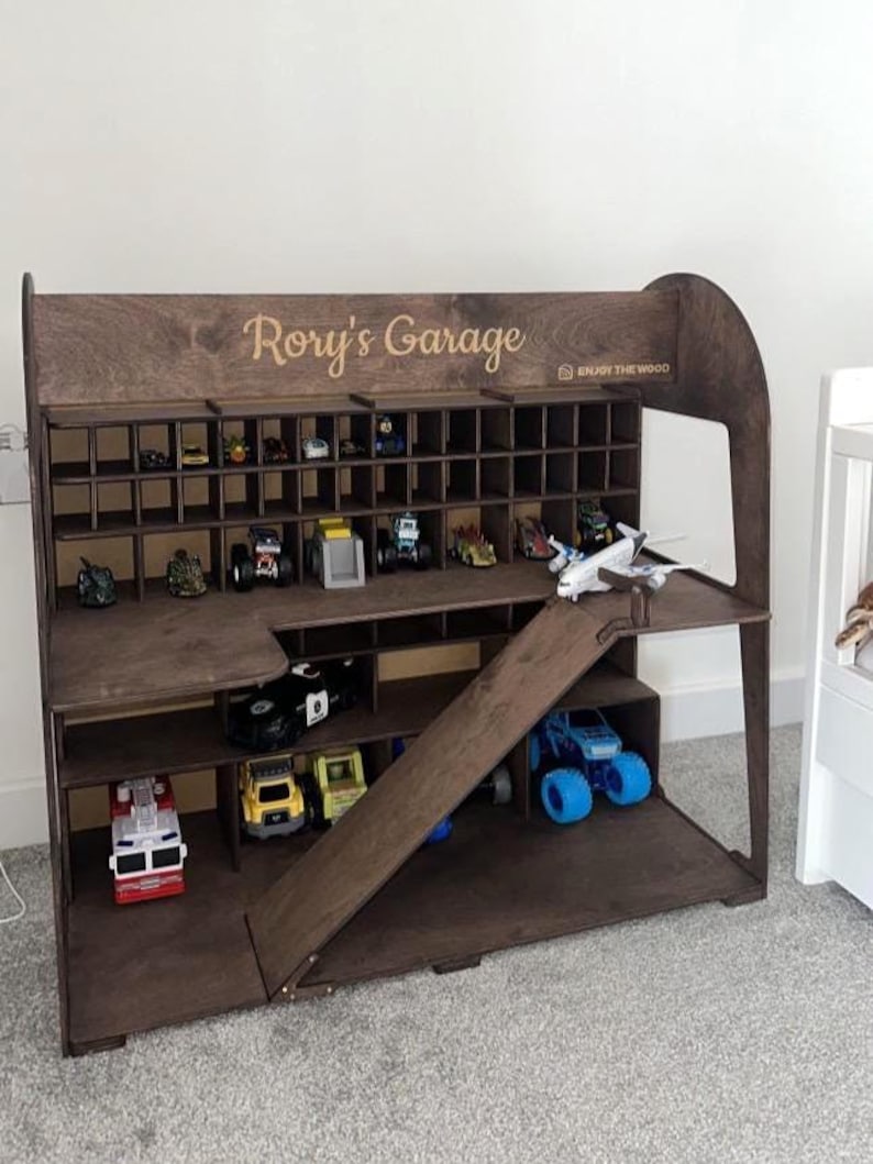 K&ouml;nnte beinhalten: Ein dunkelbraunes Spielzeug-Garage aus Holz mit mehreren F&auml;chern und einer Rampe. Es ist mit Spielzeugautos und -lastwagen gef&uuml;llt. Der Name "Rory's Garage" ist oben eingraviert. Ideal zur Aufbewahrung von Spielzeug.