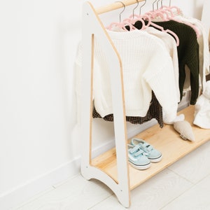 以下が含まれることがあります： 棚付きの白い木製の洋服掛け。洋服掛けには、ピンクのハンガーと白いセーター、茶色と白のチェック柄のスカート、緑のセーターがかけられています。棚には青いスニーカーが置かれています。