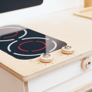 May include: A white wooden toy stove with a black stovetop and two silver knobs. The stovetop has red markings that look like heating elements.