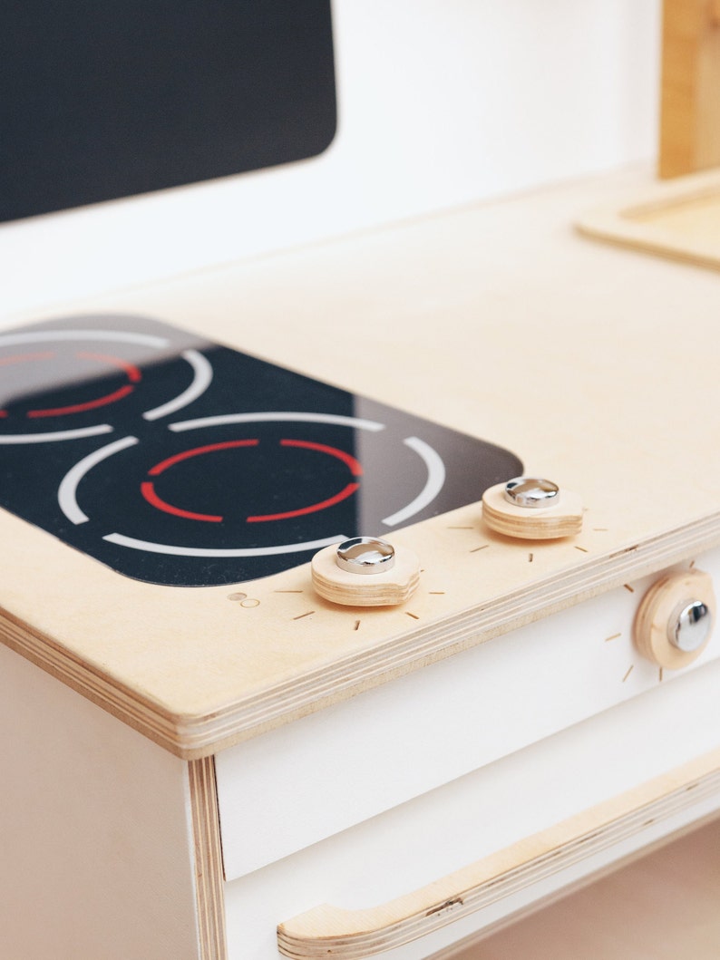 May include: A white wooden toy stove with a black stovetop and two silver knobs. The stovetop has red markings that look like heating elements.
