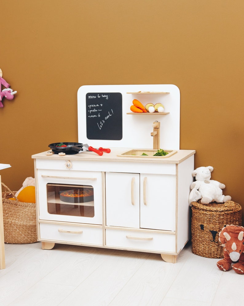 May include: White wooden play kitchen with a black chalkboard that says "menu for today: pizza, pasta, cream, let's cook!"  The kitchen has a sink, oven, and two cabinets. There are toy vegetables and a toy pizza in the oven.