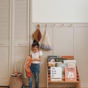 May include: A wooden bookshelf filled with children's books including titles like The Book With No Pictures and Flora Flamingo, a wicker cart with a doll, and two hanging fabric bags against a paneled wall.