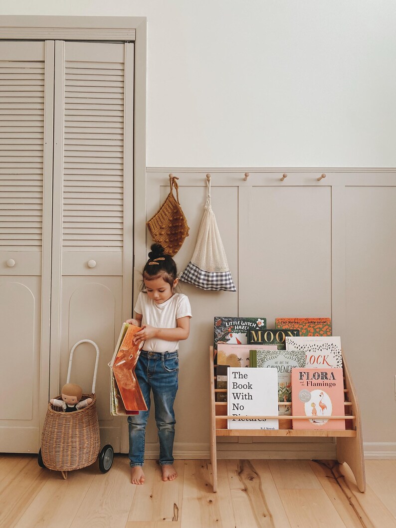 May include: A wooden bookshelf filled with children's books including titles like The Book With No Pictures and Flora Flamingo, a wicker cart with a doll, and two hanging fabric bags against a paneled wall.